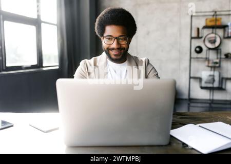 A young, stylish businessman works intently on a laptop in a well-lit modern office setting, displaying a focus and professionalism. His sharp outfit exude confidence and contemporary business attire Stock Photo
