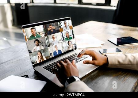Young entrepreneur is shown in the midst of a cheerful virtual meeting, with a laptop screen displaying a gallery of friendly and diverse faces. Virtual meeting and video call concept Stock Photo
