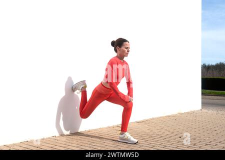 sporty woman doing leg stretching exercises outside against a white wall Stock Photo