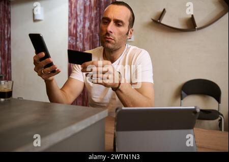 Handsome man holds credit card, using smartphone, online shopping, mobile banking. Young businessman freelancer receives payment, checks balance on mo Stock Photo