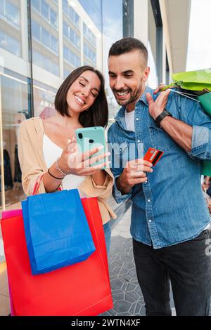 Vertical. Couple using a shopping online smartphone app to buy a sale on a mall. Young customers paying a purchase with a market place application. People using a smart phone to search a discount. High quality photo Stock Photo