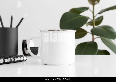 Enamel mug mockup with workspace accessories on a white table and a ficus plant. Stock Photo