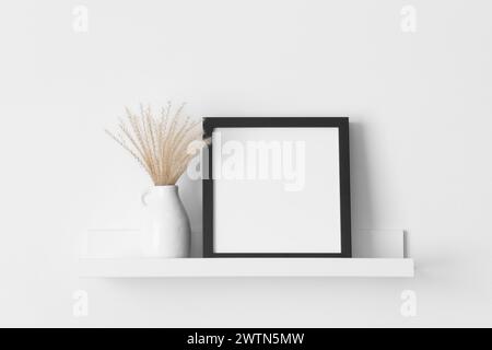 Black square frame mockup with a pampas decoration on the wall shelf. Stock Photo