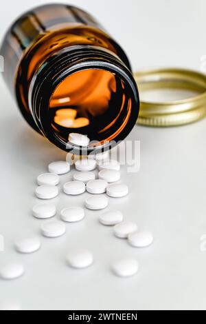 White Tablets Scattered on Table Next to Brown Glass Vial, on White Background, Close-up, Vertical Frame Stock Photo