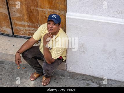 Cartagena, Colombia - July 25, 2023: young man squat on side walk against wall on Calle 30, he wears yellow shirt and blue NY Yankees cap Stock Photo