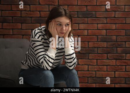 Sad young woman sitting on chair near brick wall, space for text Stock Photo