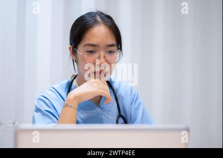 A professional and serious Asian female doctor is reading medical cases on her laptop, working in her office at the hospital. Stock Photo
