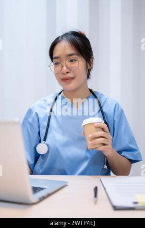A professional and experienced Asian female doctor is sipping coffee while reading medical cases on laptop in her office. Stock Photo
