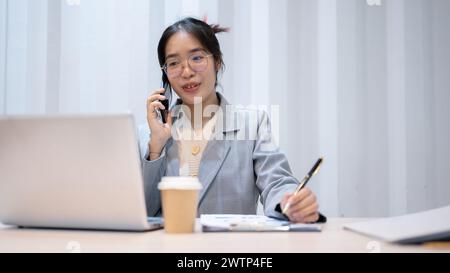 A confident Asian businesswoman is multitasking, talking on the phone with her client while working in her office. Stock Photo