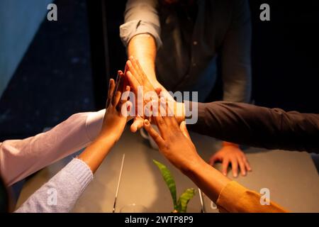 The image displays a group of hands from individuals of varying ethnicities engaging in a collective high-five above a meeting table, highlighting a moment of team success or agreement within a business context. The diversity of the team is represented by the different skin tones and the inclusivity of their workplace culture. The warm lighting focuses on their connected hands, which are at the center of the composition, symbolizing unity and shared accomplishment. Collaborative High Five in a Business Setting. High quality photo Stock Photo