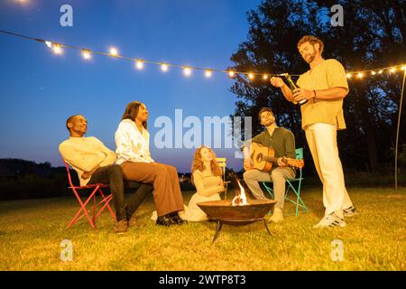 In the tranquil evening air, a group of friends relaxes around a crackling fire pit, with one playing the guitar, all under the gentle twinkle of string lights in the background. Friends Enjoying a Fireside Guitar Session Under String Lights. High quality photo Stock Photo