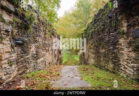A concrete path runs between two high, rough stone walls streaked with moss and rust toward autumn trees at Harpers Ferry National Historical Park. Stock Photo