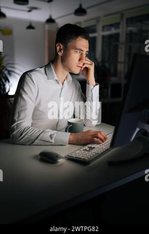 Vertical portrait of IT programmer checking computer code using desktop. Busy developer working late writing software language in dark office. Stock Photo