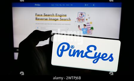 Person holding mobile phone with logo of facial recognition search company PimEyes in front of business web page. Focus on phone display. Stock Photo