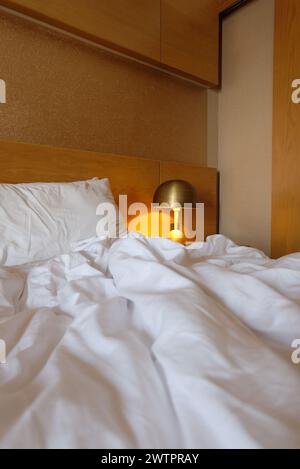 Selective focused of messy bedroom with lamp near bed. Wrinkled bed sheets and pillows. Messy bedroom, white pillow with blanket lying on bed unmade. Stock Photo