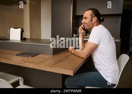 Young Caucasian man smiling while talking on mobile phone over cup of freshly brewed espresso coffee. Happy Caucasian guy receiving call from friends, Stock Photo