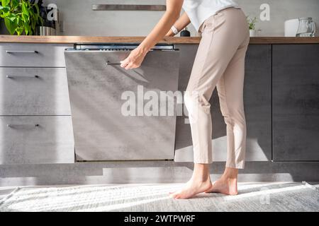 Young woman turns on the dishwasher by pressing buttons on the panel. Household and useful technology concept.  Stock Photo