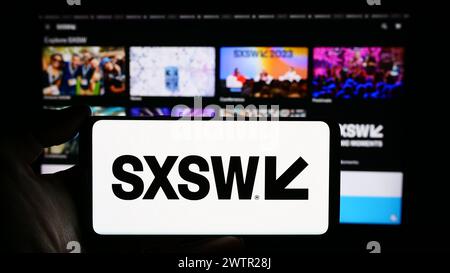 Person holding cellphone with logo of annual film and media event South by Southwest (SXSW) in front of webpage. Focus on phone display. Stock Photo