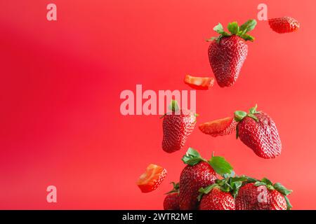 Whole and cut strawberries falling on a pile of other strawberries. isolated on vibrant red background. Stock Photo