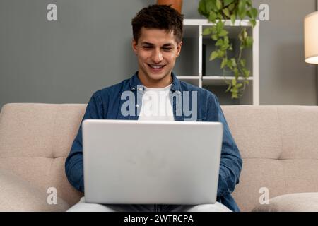 Handsome relaxed male student working with laptop while sitting on the couch. Smiling young carefree man using computer at home. Stock Photo