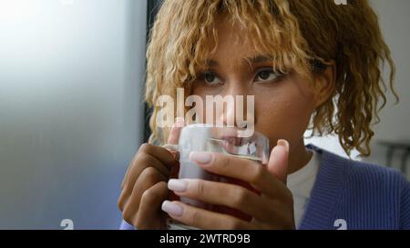 Ethnic African American happy smiling inspired female at home drinking hot herbal healthy taste black tea looking at window woman girl holding cup Stock Photo