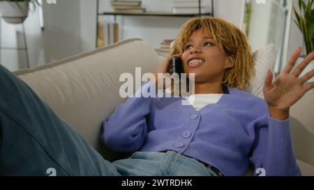 African American woman ethnic girl happy relax on couch lying on sofa at home relaxing talking mobile phone call friendly distant communication Stock Photo