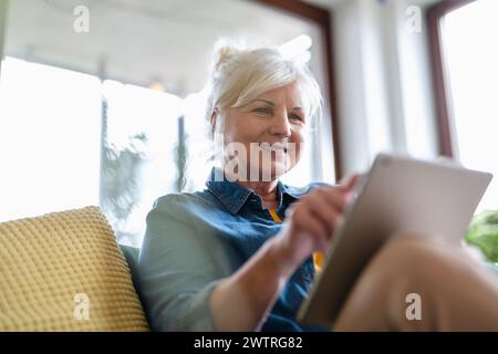Mature woman using digital tablet while sitting on sofa at home Stock Photo