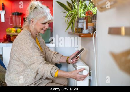 Senior woman holding mobile phone while adjusting the thermostat on the radiator at home Stock Photo