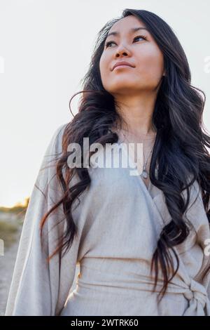 Young attractive Asian with long wavy hair outdoors. A charming Korean woman in a linen dress enjoys nature. A beautiful girl looks into the distance Stock Photo