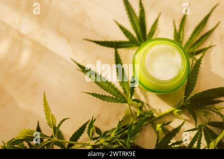 A layout of a jar of cream with cannabis leaves on a marble table. A mock-up of a glass bottle with natural white cream and hemp on a beige background Stock Photo