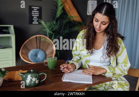 Charming curly-haired brunette in casual clothes signs documents at home. Young attractive woman fills out paper forms in living room. Businesswoman r Stock Photo