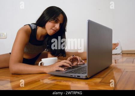young latin brunette woman at home lying on floor calm and quit, studying remotely with laptop, typing on computer keyboard and watching screen Stock Photo