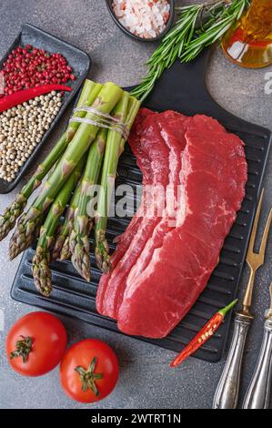 marbled beef bacon. black wooden cutting board with raw marbled beef bacon with asparagus, seasoning tomatoes and assorted peppers. flat lay on gray b Stock Photo