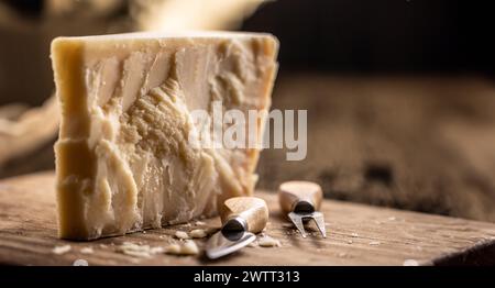 A block and pieces of Parmesan cheese on a cutting board along with a chisel and a skewer. Stock Photo