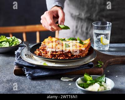 A person garnishing a freshly baked lasagna with basil leaves on a rustic table setting. Stock Photo