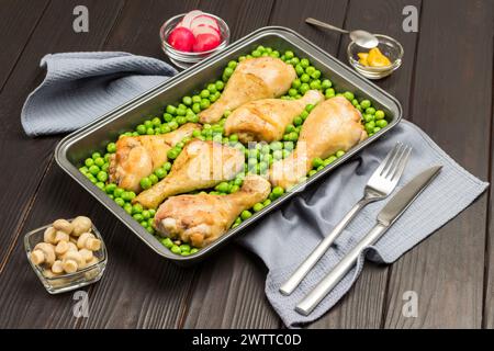 Chicken drumsticks and green peas in metal baking tray. Mustard, radish on table. Fork and knife on gray napkin. Dark wooden background. Top view Stock Photo