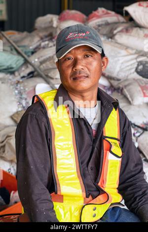 Young worker in reflective vest sitting amidst industrial materials in a plastic recycling factory, Thailand Stock Photo