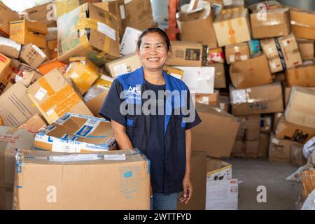 A smiling worker in a recycling factory surrounded by piles of parcels, Thailand Stock Photo