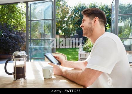 Man smiling while using a smartphone at a sunny kitchen table Stock Photo