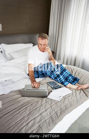 Man working on bed with a laptop and paperwork Stock Photo