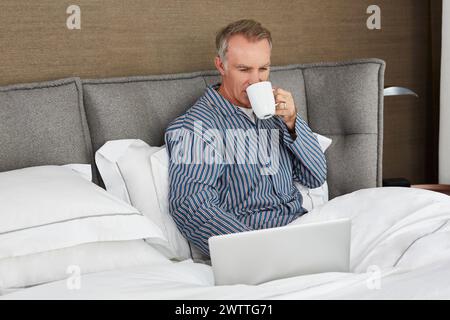 Man working on laptop in bed while drinking coffee Stock Photo