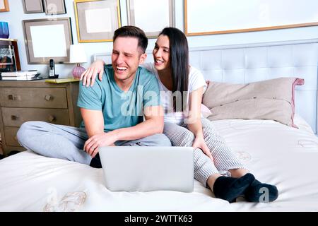 Couple smiling and using a laptop on the bed. Stock Photo