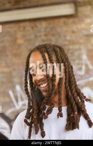 Smiling man with dreadlocks standing against a brick wall Stock Photo