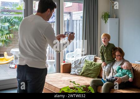 Family moment captured indoors as a man takes a photo of a woman with two young children on a cozy sofa. Stock Photo