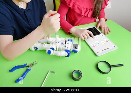 Children's hands assemble robot on workbench, screwing parts while reviewing programming code on tablet. Concept of learning and hands-on education Stock Photo