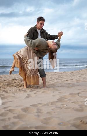 Man twirling a woman in a flowing dress on the beach, both in mid-laugh, a weeding or engagement dance. A lively, joyful moment, great for lifestyle c Stock Photo