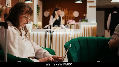 Senior couple passing time in lounge area, woman using laptop to create holiday itinerary while man enjoys lecture on couch. Retired people waiting to see accommodation at hotel. Stock Photo