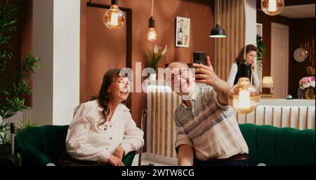 Senior couple talk on online videocall with nephews while travelling abroad, internet videoconference connection to present vacation plans. People passing time in hotel lounge area. Stock Photo