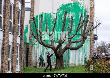 London, UK. 19th March, 2024. A member of the public interacts with new artwork by world-renowned street artist Banksy which had appeared the previous day on the side of a residential building behind a tree located in the garden of a social housing block in Finsbury Park. The artwork, claimed by the street artist, takes the form of green paint sprayed onto a wall behind a pollarded cherry tree so as to appear like foliage when viewed from the front, together with a stencil of a woman holding a pressure washer. The shade of green paint used appears to match that used by Islington Council for it Stock Photo