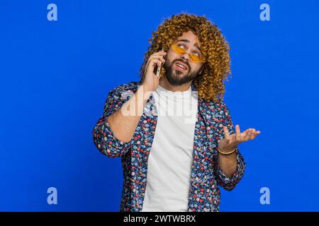Disinterested man with curly hairstyle wig having annoyed boring talk on smartphone. Guy tired sleepy guy talking on mobile phone with friend, making online conversation on blue background. Copy-space Stock Photo
